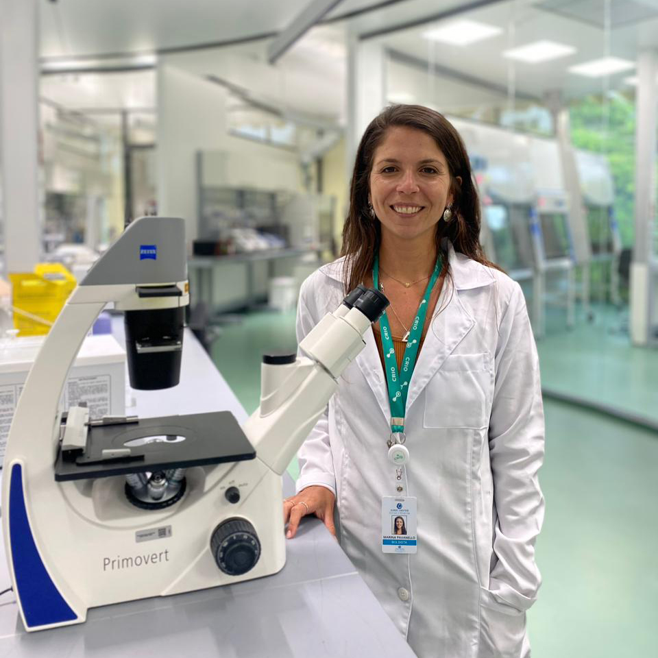 A Imagem mostra a pós-doutoranda do centro de Pesquisa em Imuno-oncologia (CRIO), Marina Pavanello, em laboratório vestindo jaleco, ao lado de um equipamento que parece ser um microscópio.