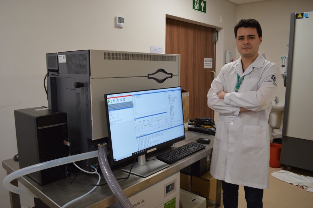 Homem branco de jaleco branco e de cabelo curto castanho escuro. Ele está de braços cruzados, posando ao lado de uma máquina chamada "espectrômetro de massas" que serve para ajudar na detecção de cânceres. Na imagem há um monitor de computador em que exibe um gráfico que revela dados de detecção de massas.